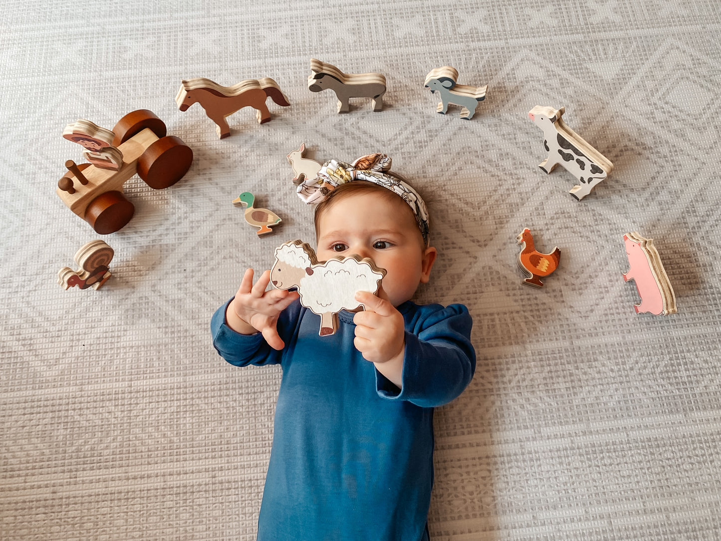 A baby wearing a blue outfit and a patterned headband lies on a textured light gray blanket, holding a wooden animal toy while surrounded by other wooden farm and animal toys.