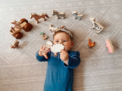 A baby wearing a blue outfit and a patterned headband lies on a textured light gray blanket, holding a wooden animal toy while surrounded by other wooden farm and animal toys.