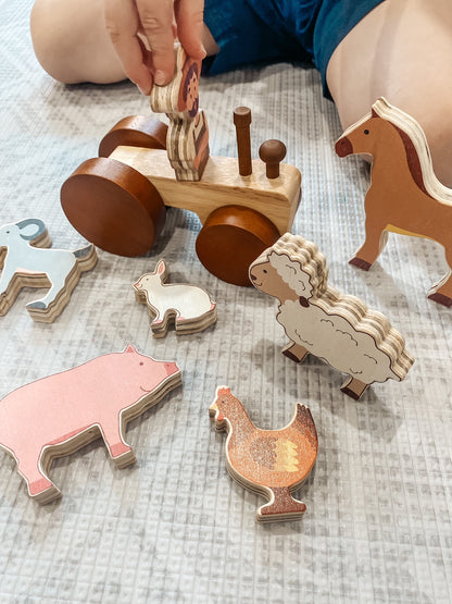 A child's hand is playing with a wooden farm animal toy set on a textured light gray blanket. The set includes a wooden tractor with round wheels and handle, several animal cutouts (pig, chicken, sheep, rabbit, horse), and wooden pegs.