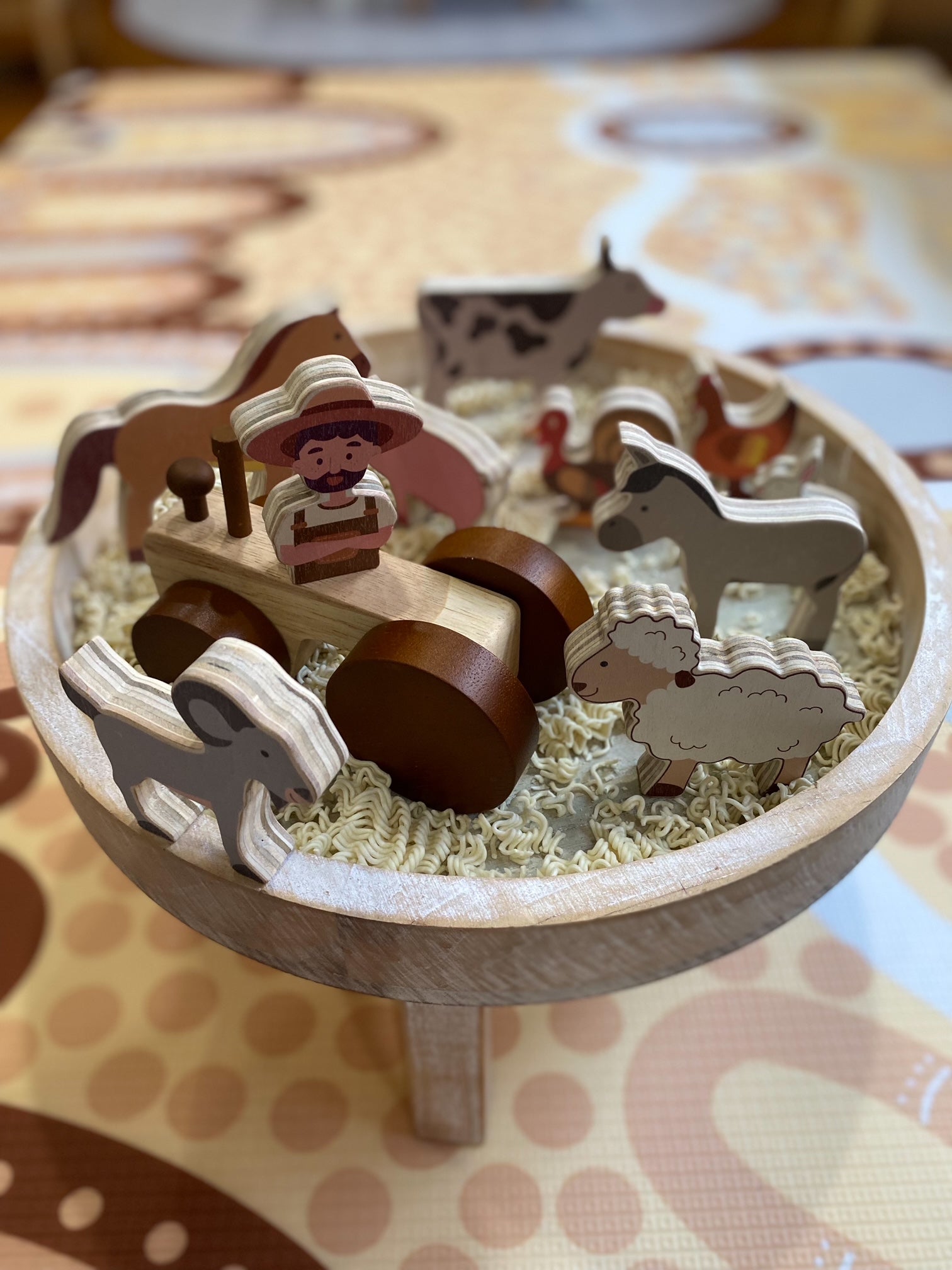 A wooden farm-themed toy set displayed in a shallow wooden bowl filled with shredded paper. The set includes a small tractor with a driver figure, several horses, a cow, a sheep, and a chicken, all made from light and dark natural wood.