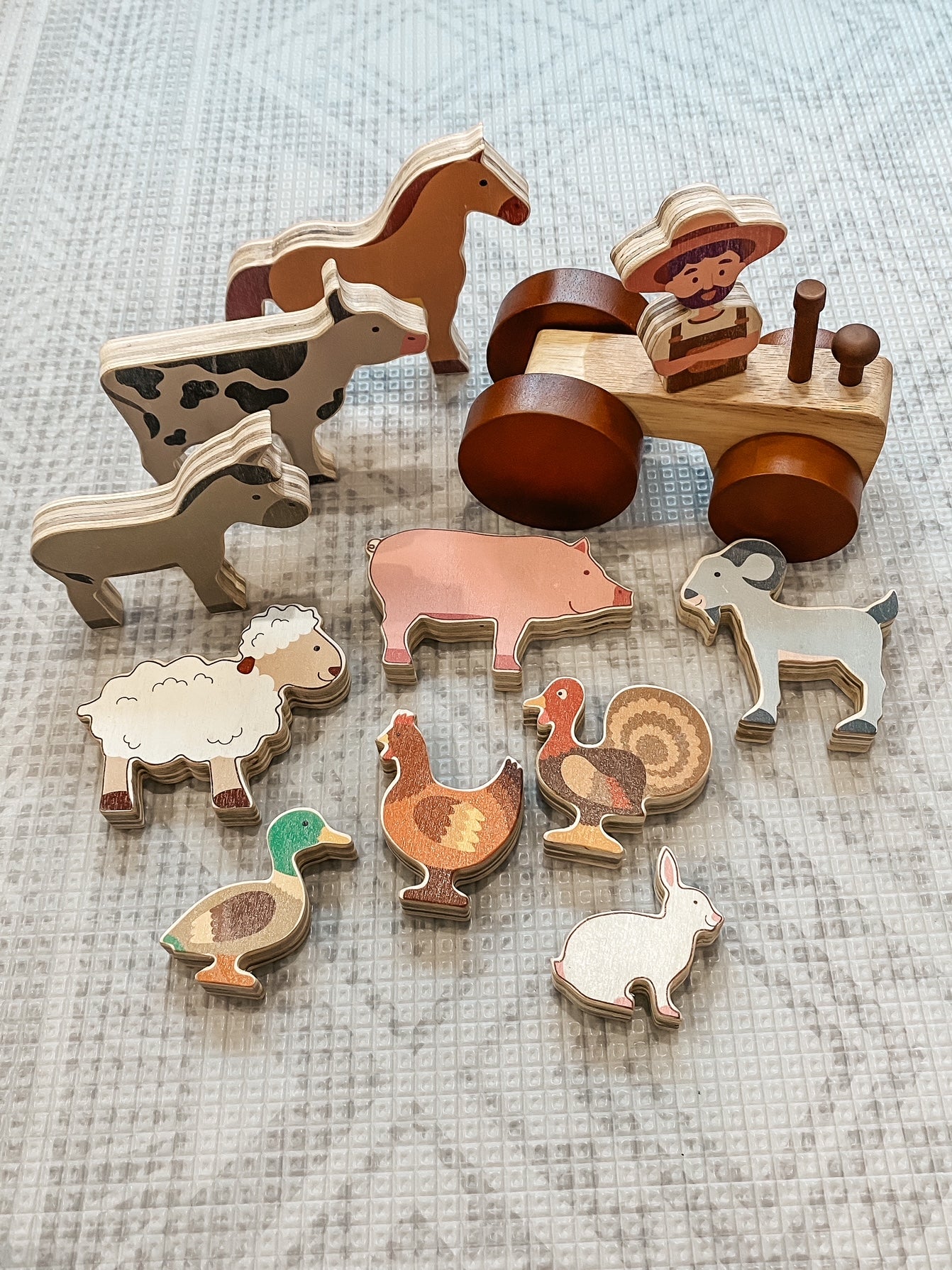 A set of wooden farm-themed toy pieces arranged on a light textured fabric background, including animal figures and a tractor with a driver.