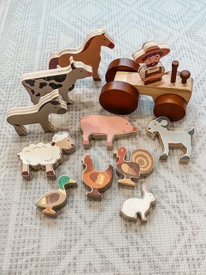 A set of wooden farm-themed toy pieces arranged on a light textured fabric background, including animal figures and a tractor with a driver.