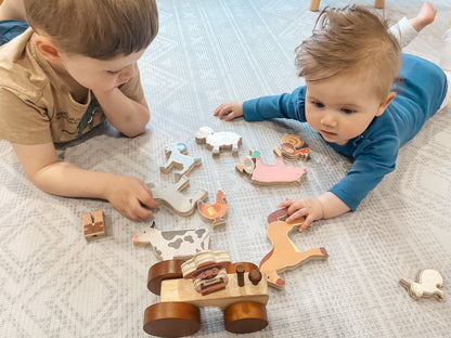 Two toddlers lying on a light patterned play mat, playing with a wooden farm animal puzzle set. The set includes wooden animal pieces with painted accents such as a cow, horse, chicken, and a small cart.