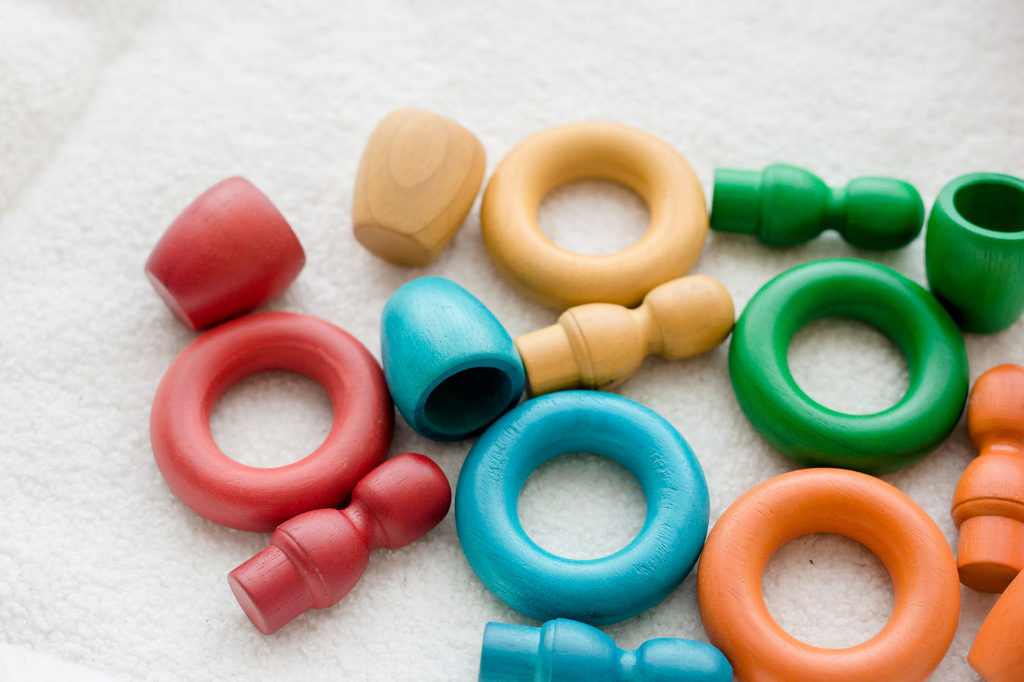 A collection of colorful wooden baby teething toys and rings in red, blue, green, orange, and natural wood, scattered on a soft white textured fabric background.