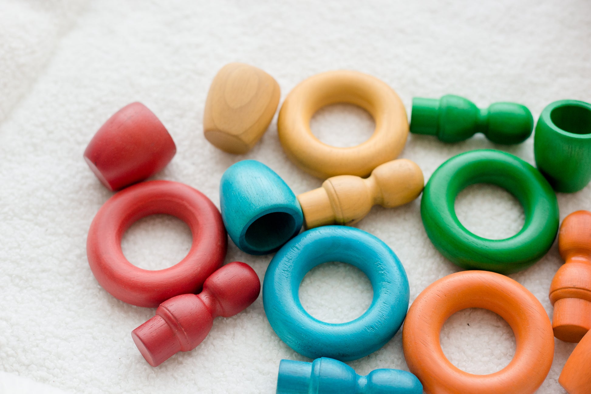 A collection of colorful wooden baby teething toys and rings in red, blue, green, orange, and natural wood, scattered on a soft white textured fabric background.