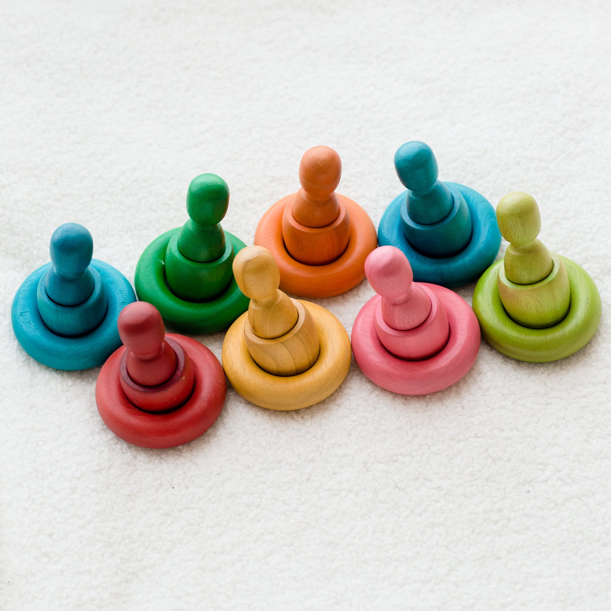 A set of colorful wooden stacking rings and pegs for baby or toddler sensory play, arranged on a soft white textured fabric background.