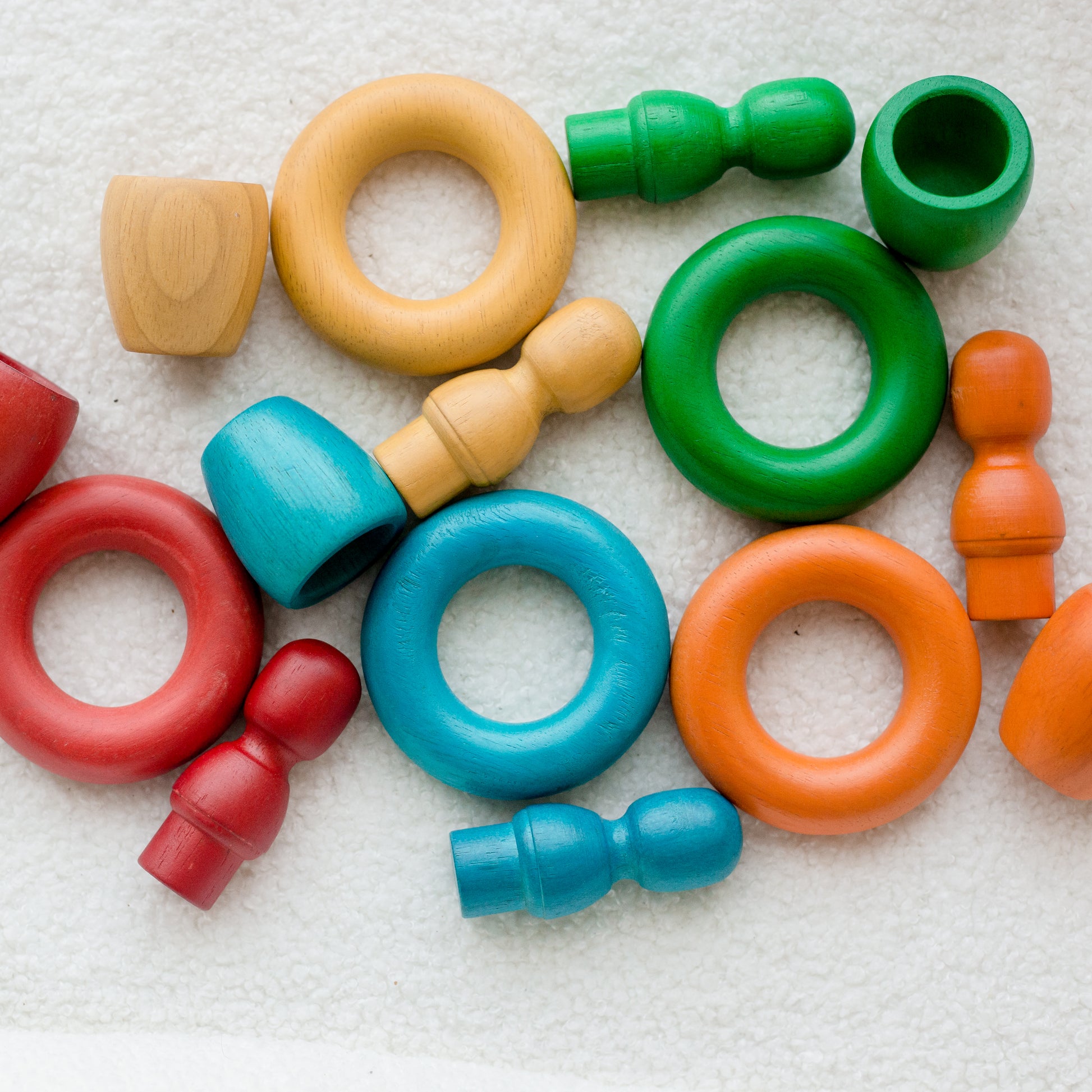 A set of colorful  wooden rings in red, blue, green, orange, and natural wood, arranged on a textured white fabric background.