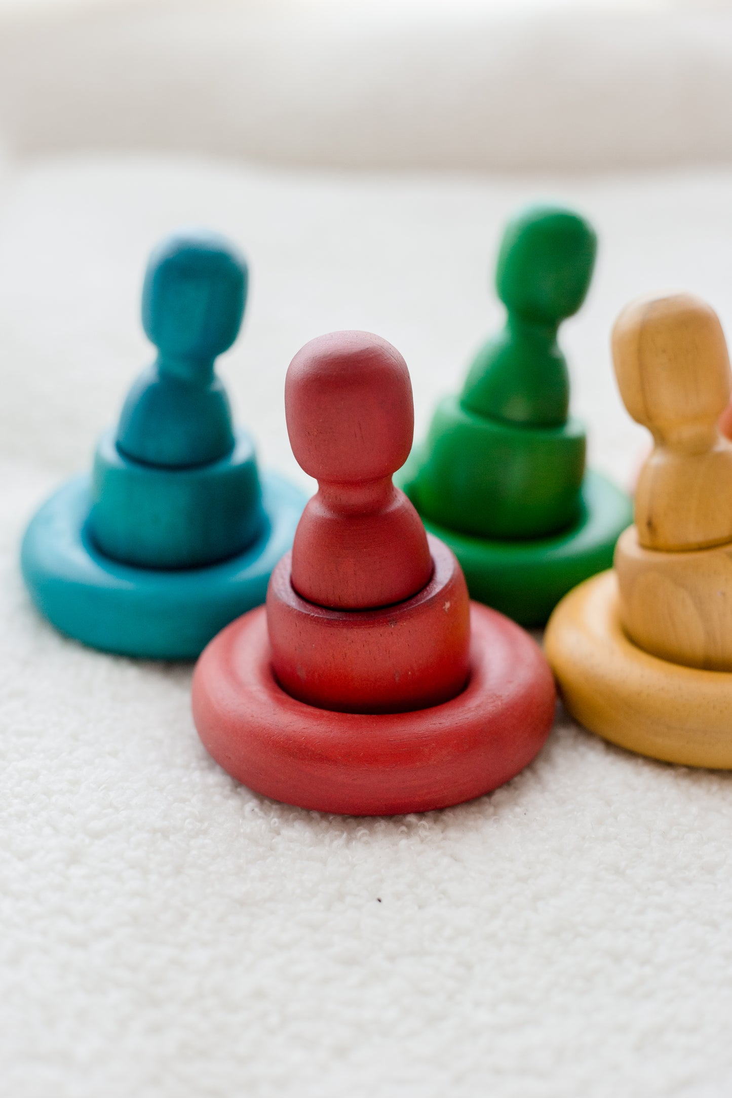Four colorful wooden peg and ring stacking toys arranged on a soft white textured surface. The set includes teal, red, green, and natural wood versions.