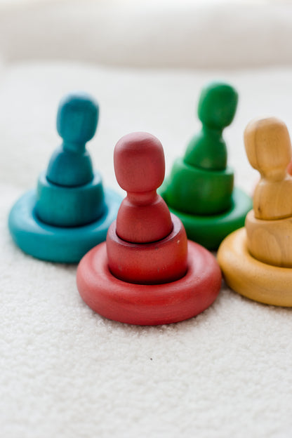 Four colorful wooden peg and ring stacking toys arranged on a soft white textured surface. The set includes teal, red, green, and natural wood versions.